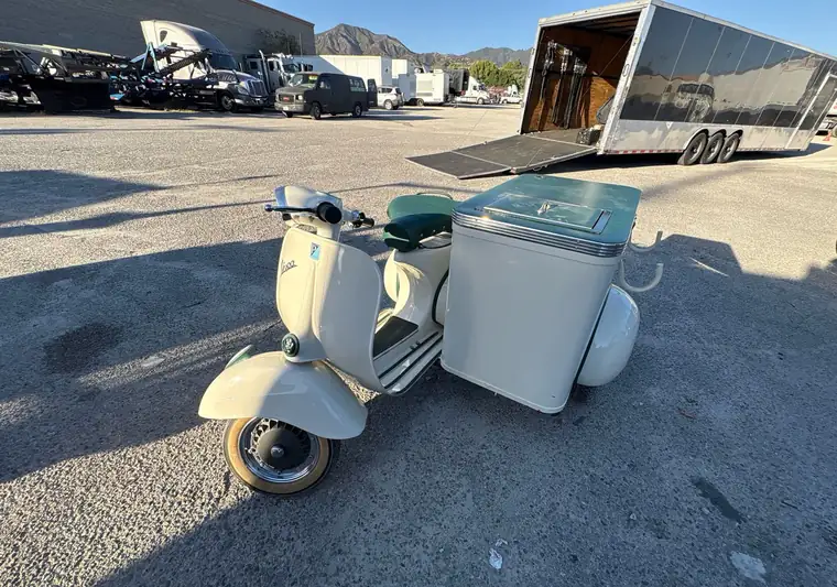 Cream 1965 Vespa with a sidecar in a yard beside an enclosed race-style trailer with its ramp lowered, at pickup in Los Angeles, California